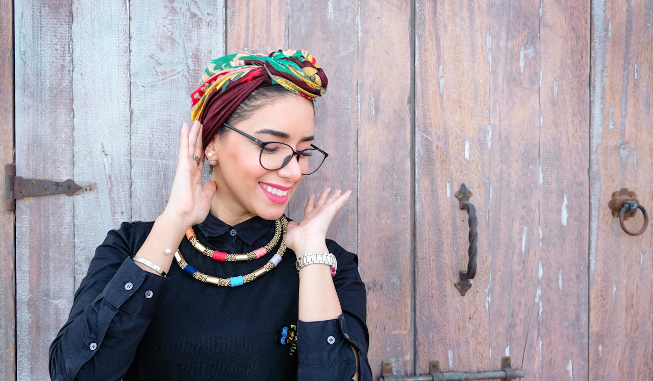 A fashionable woman with a headscarf and necklace smiling in front of a rustic wooden door.